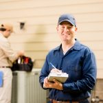 Smiling HVAC technician with another technician in the background tending to outdoor AC unit